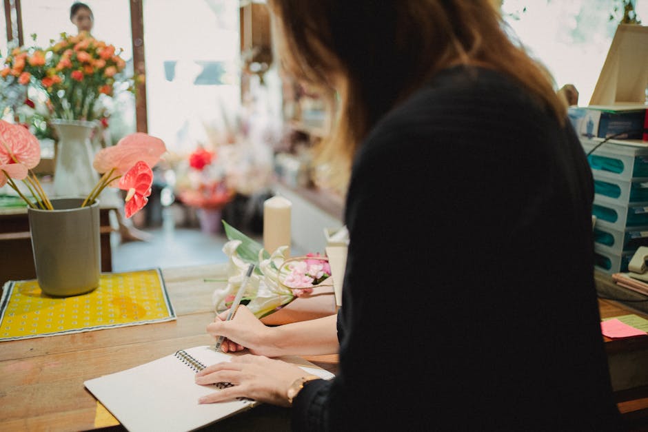 A florist at Millbank Florist, captured in a close-up view, is carefully preparing a floral arrangement on a wooden worktable. She is wearing a dark top and has shoulder-length, slightly wavy brown ha